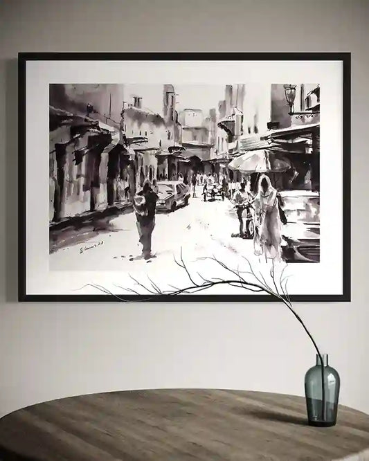 Framed black and white street scene artwork by Elamin Awad hanging above wooden table with glass vase and branch