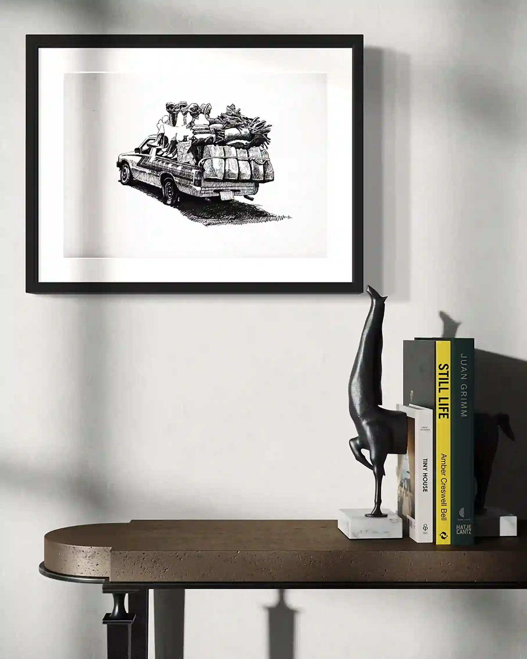 Framed Long Road ink artwork by Obada Gabir displayed on wall above wooden shelf with sculpture and books