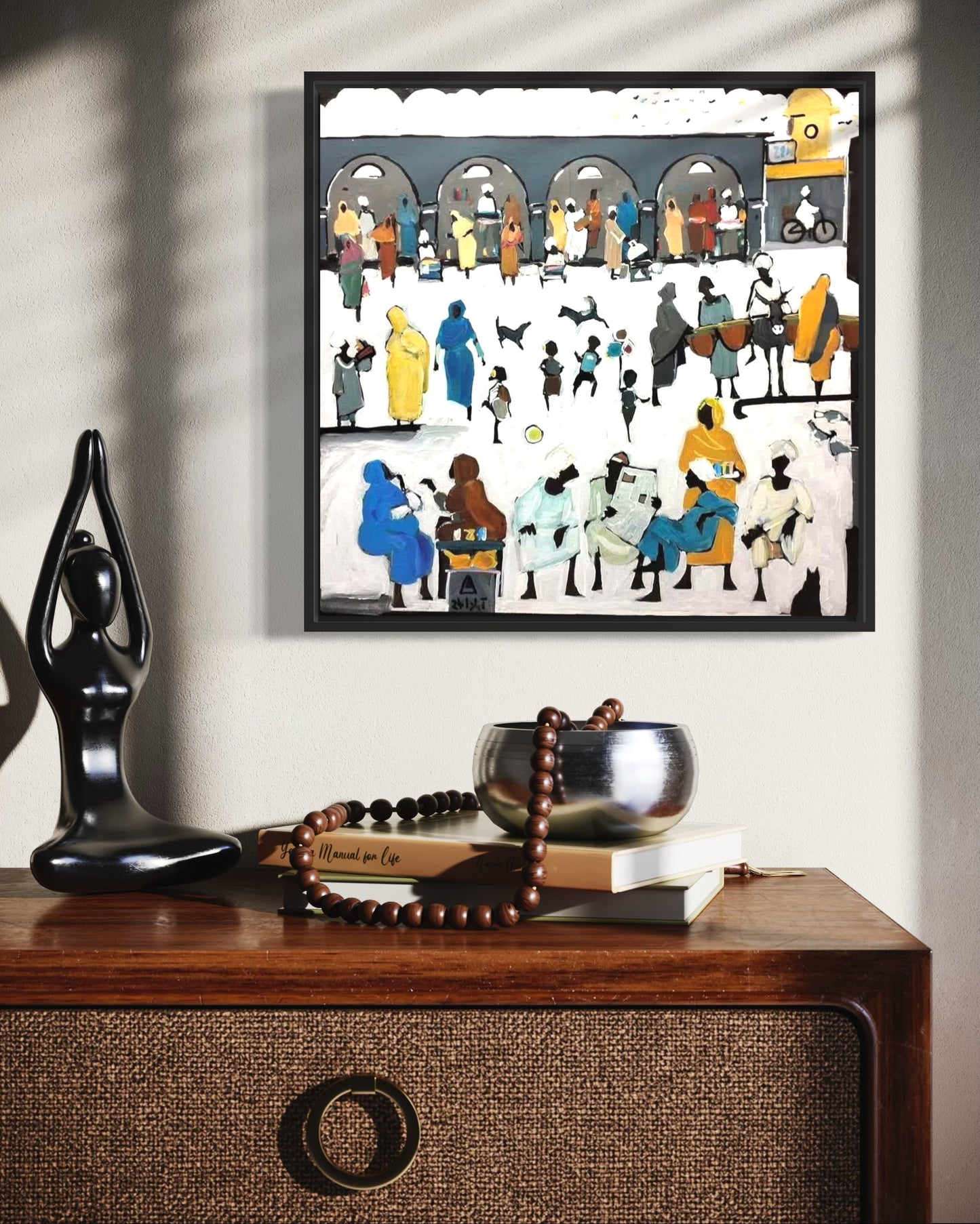 Framed artwork of people in a market scene on a wall above a wooden cabinet with decorative items.