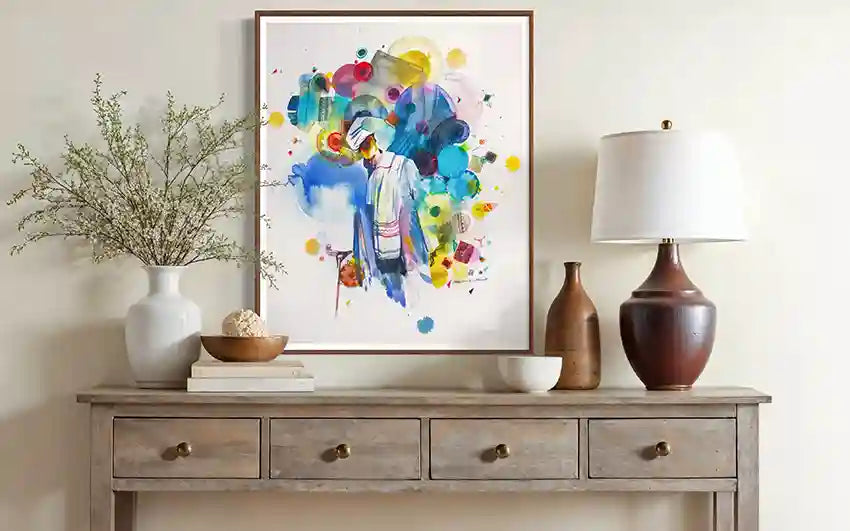 Decorative console table with a colorful abstract art print by artist Galal Yousif, lamp, and vases against a white wall.