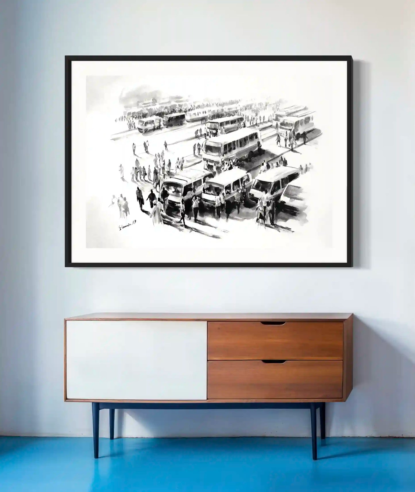 Framed black and white bus station artwork by Elamin Awad hanging above mid-century modern wooden credenza on blue floor